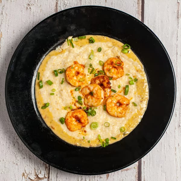 Shrimp and grits with green onions and seasoning in a black bowl on a white wooden surface.