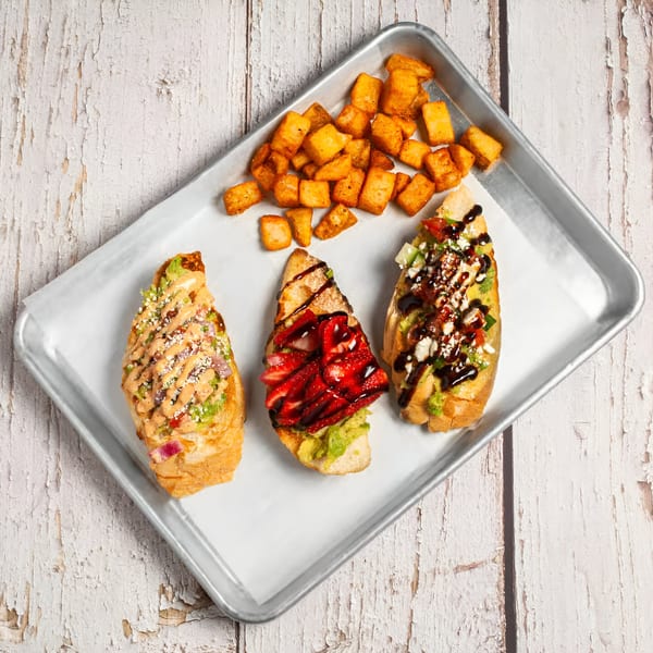 Three True Grit American Bistro breakfast bruschettas with various toppings and a side of crispy potato cubes.
