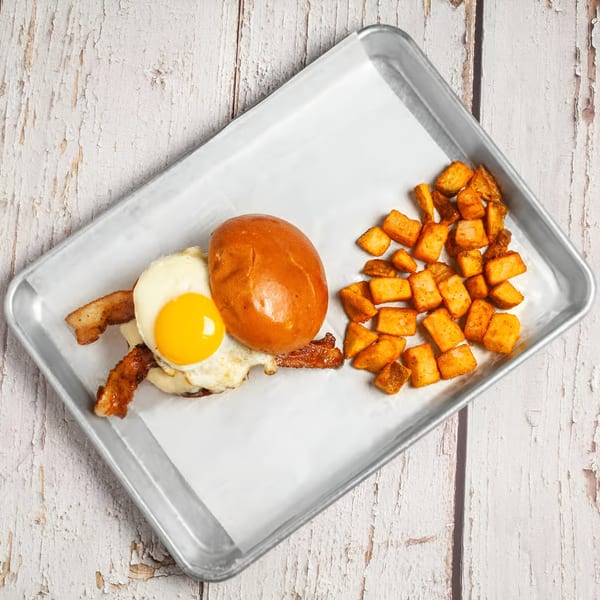True Grit American Bistro's breakfast sandwich with fried egg, bacon, and home fries on a metal tray.