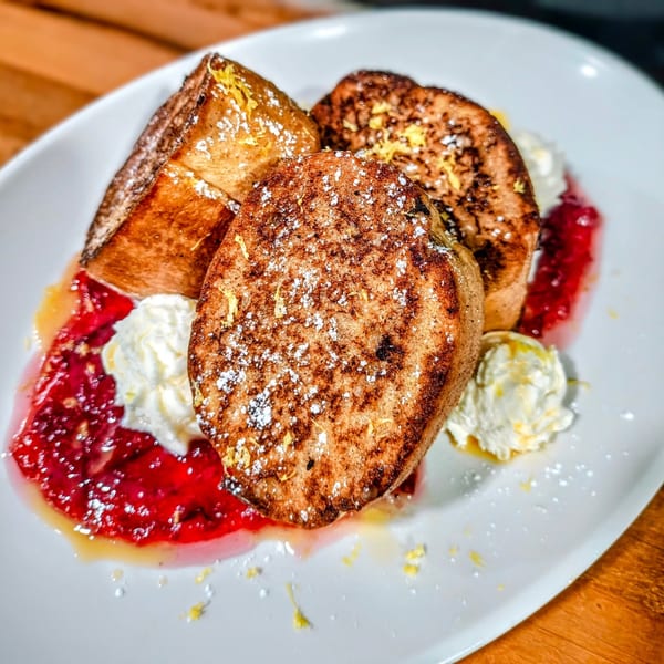 Thick-cut French toast with powdered sugar, lemon zest, raspberry sauce, and whipped cream.
